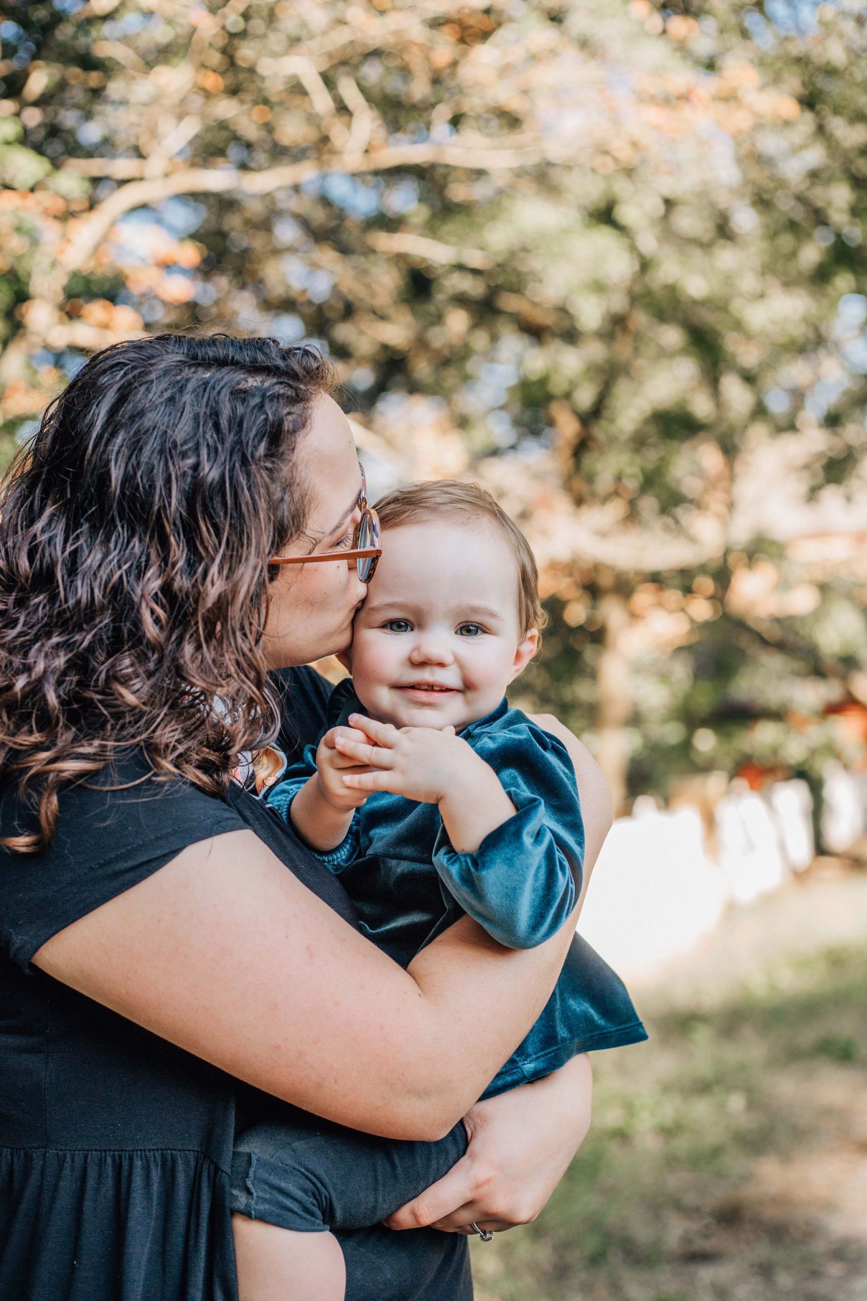 mom holding her toddler and kissing her on the head, as the toddler smiles at the camera for fall family photos in Hampton Roads, blog post inspiration to plan family photo outfits