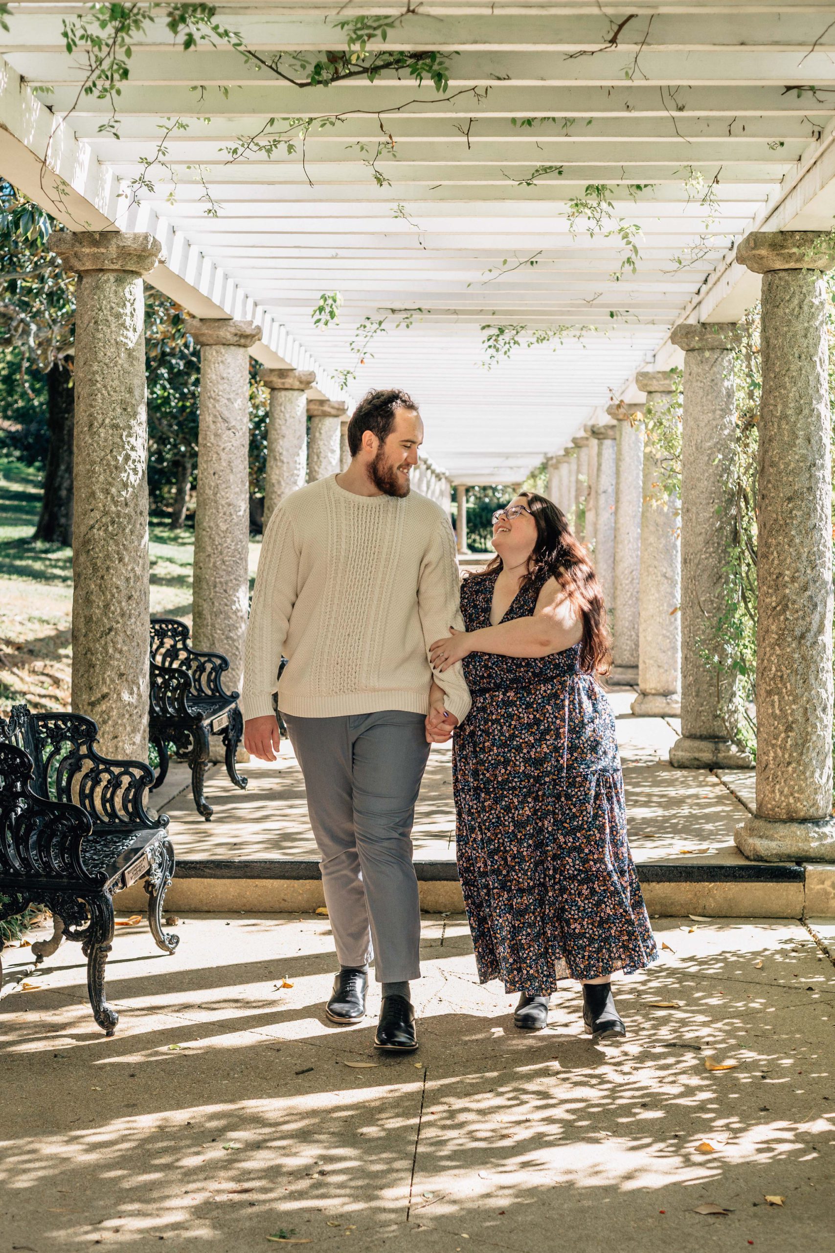 man and woman holding hands and walking to the camera under a pergola with greenery growing around them for their summer Hampton Roads Natural Engagement Photo Pose
