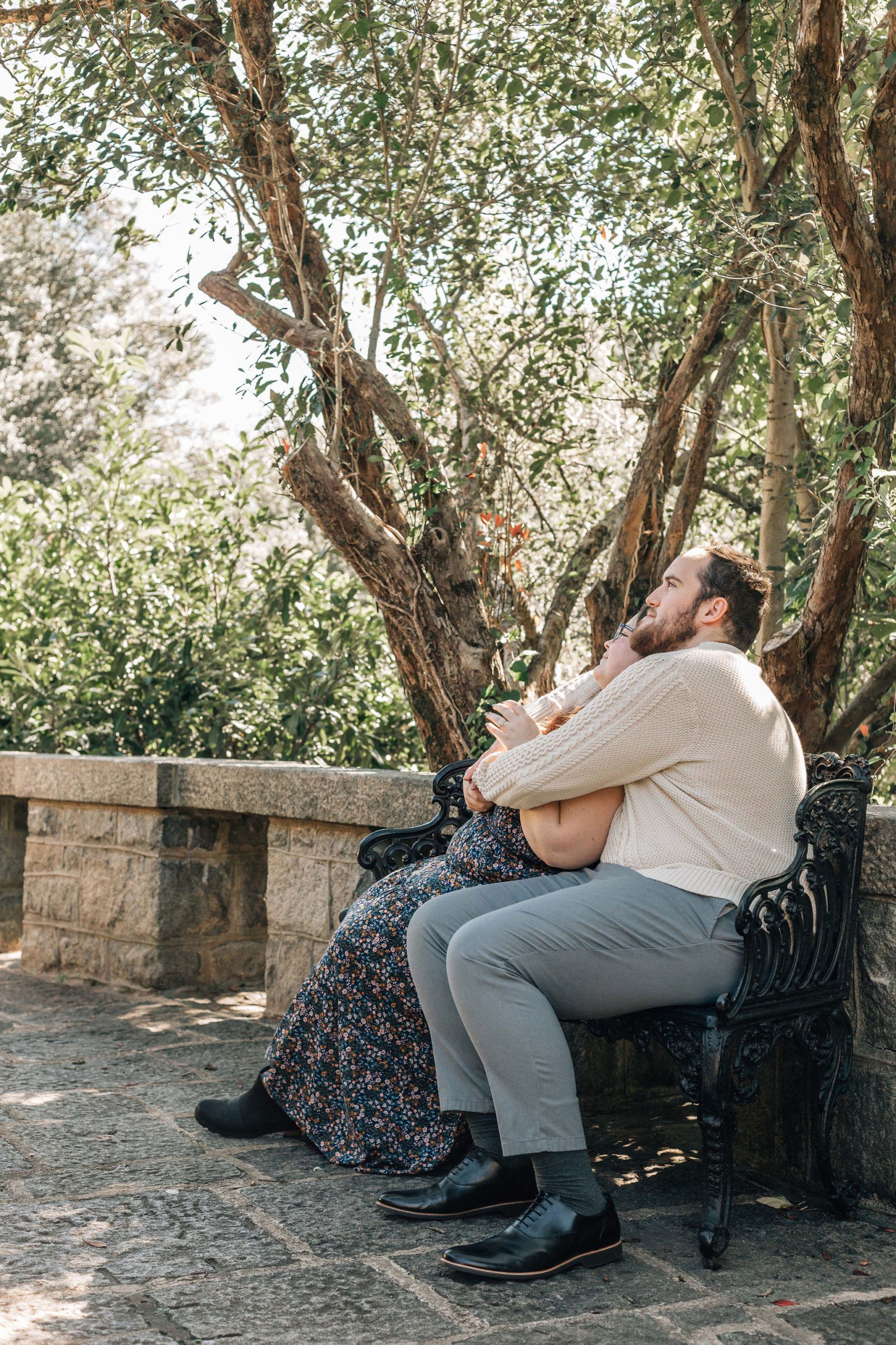 man and woman sitting on a park bench together and cuddling as they look up in the trees Natural Engagement Photo Pose
