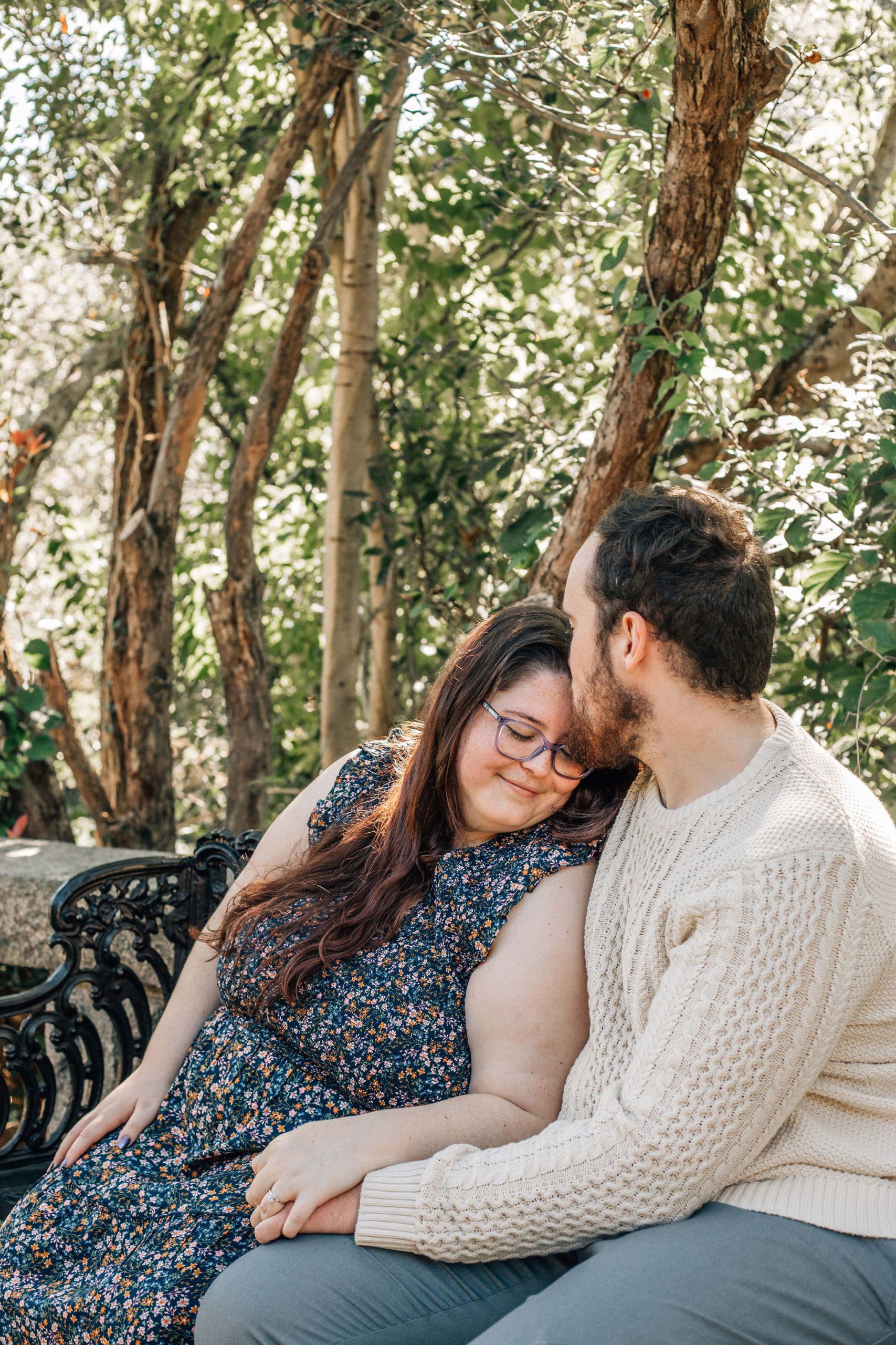 man leaning into his fiance as they sit on a park bench in Virginia as he kisses her forehead and she lightly smiles at wedding garden venue maymont park natural engagement photo pose ideas