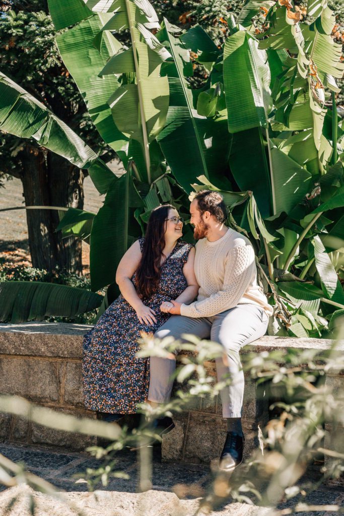 man and woman sitting in a garden together in Hampton Roads for their engagement photos in the summer as they smile at one another talking about the maymont park wedding venue