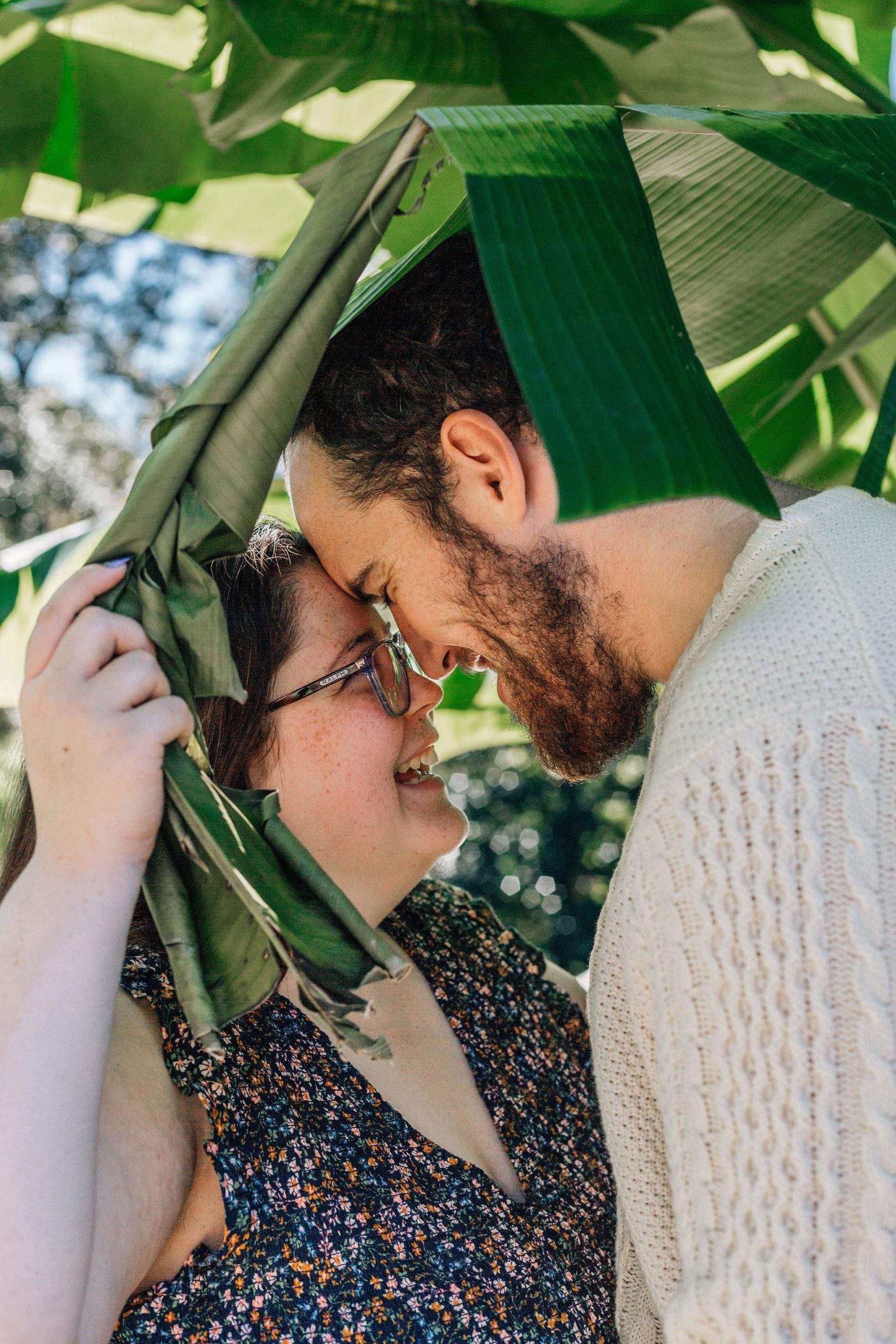 Hampton Roads engagement photographer capture man and woman standing under a tree with large leave as they laugh together for their engagement photos and natural-engagement-photo-ideas