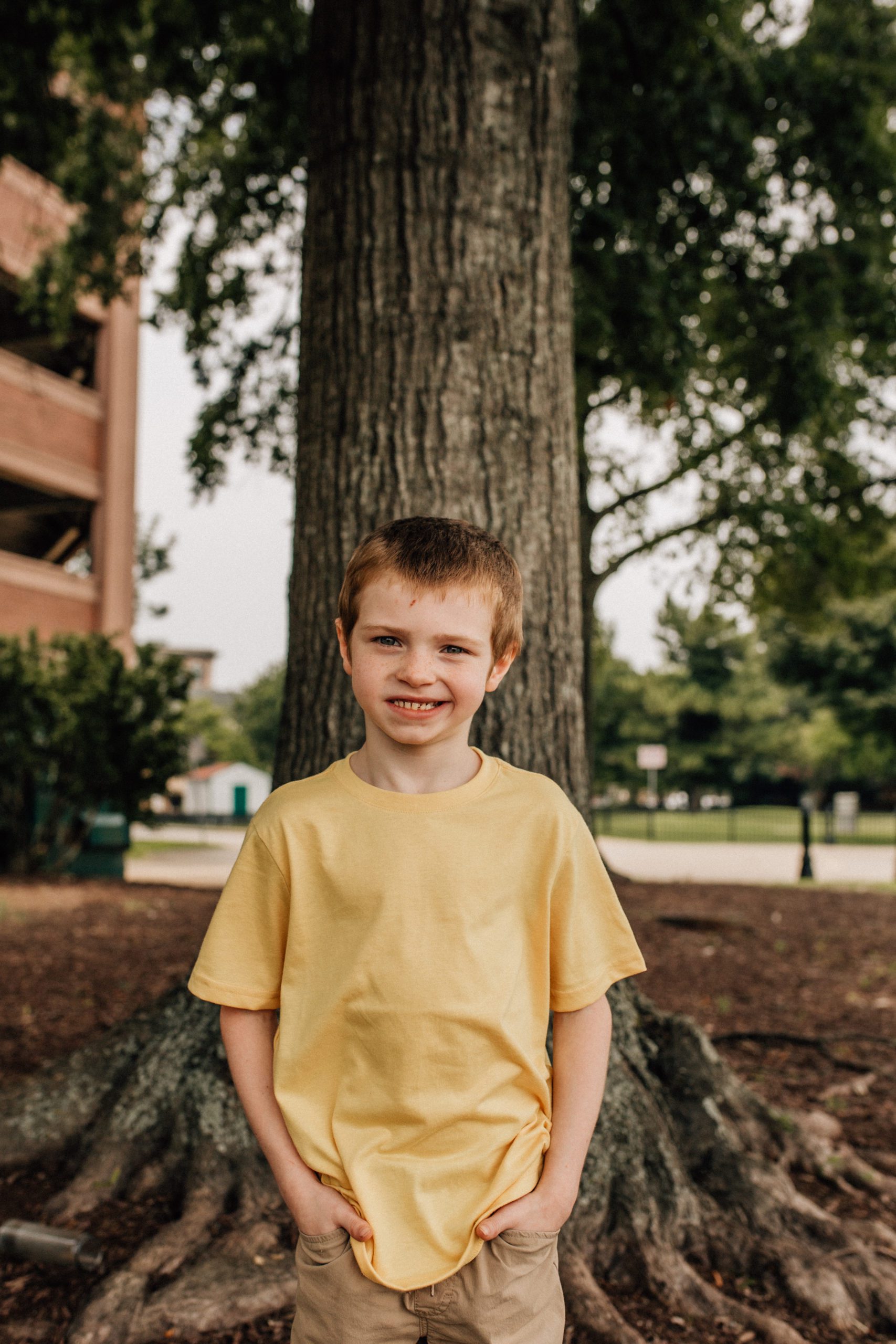 little boy with his hands in his pockets smiling at the camera while standing in front of a tree captured by Hampton Roads family photographer