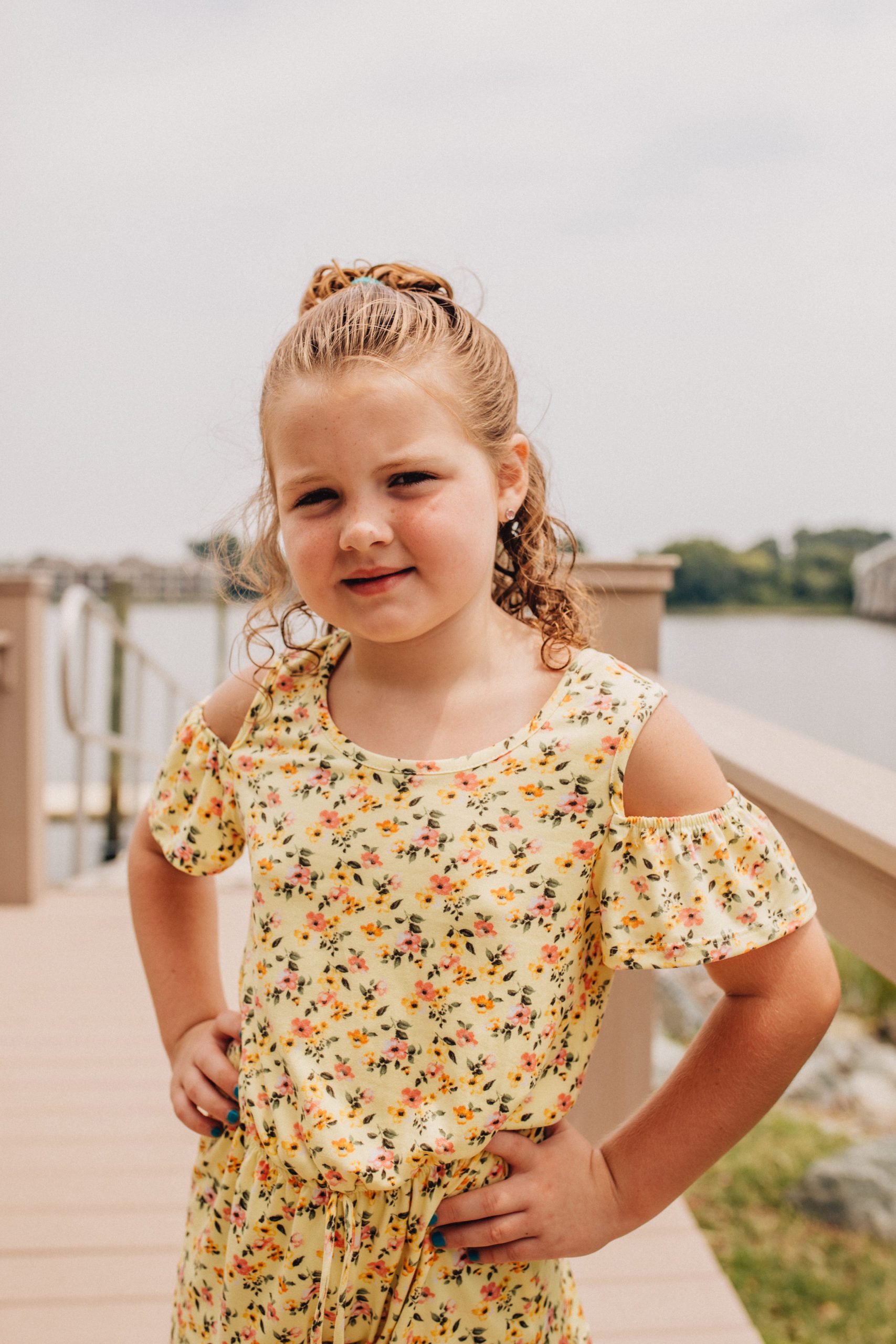 daughter posing with her hands on her hips and smiling at the camera with the water behind her as she wears a yellow dress for Hampton Roads family photos with young children