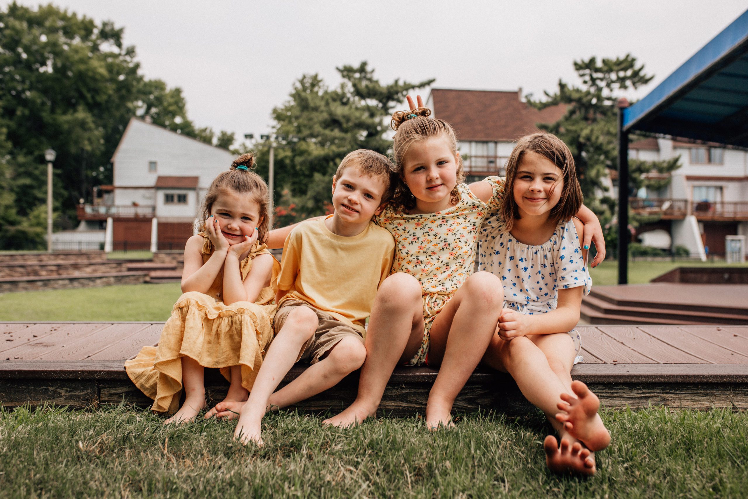 four little siblings sitting on a curb together at a park with their arms around each others shoulders for Hampton Roads family photography