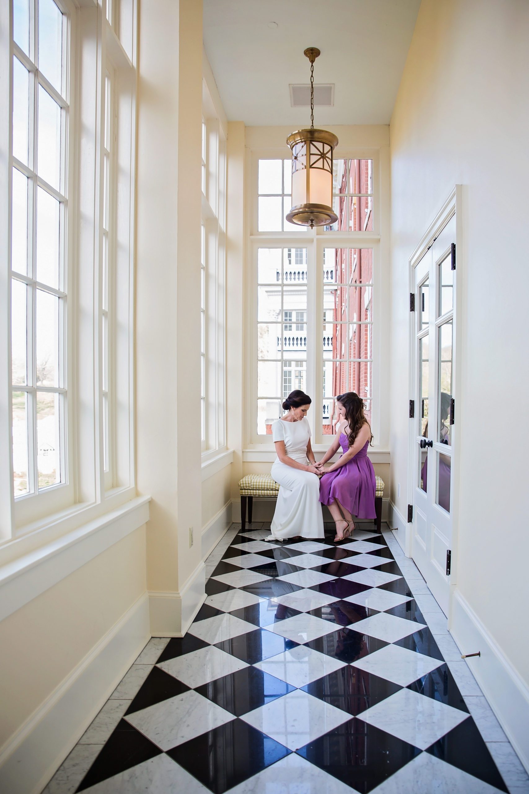 bride sitting with her maid of honor in a long hall as they hold hands and talk